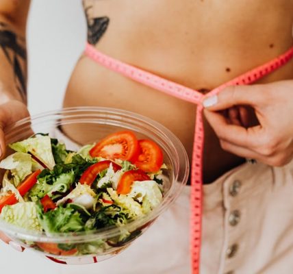 Woman holding vegetable salad and measuring waist with tape, symbolizing healthy lifestyle choices.