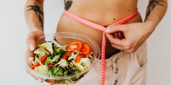Woman holding vegetable salad and measuring waist with tape, symbolizing healthy lifestyle choices.